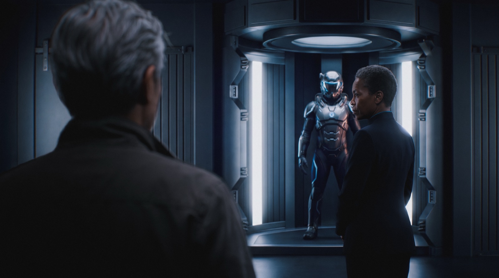 Kai facing the illuminated exo-suit in its chamber
